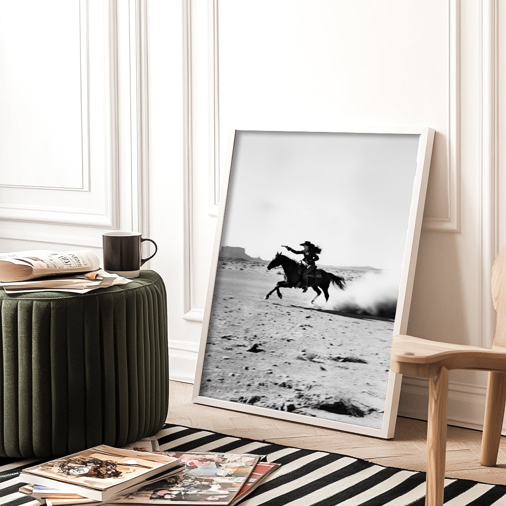 A black-and-white photograph of a cowboy galloping on horseback through the desert, with dramatic light and shadow across the sand.