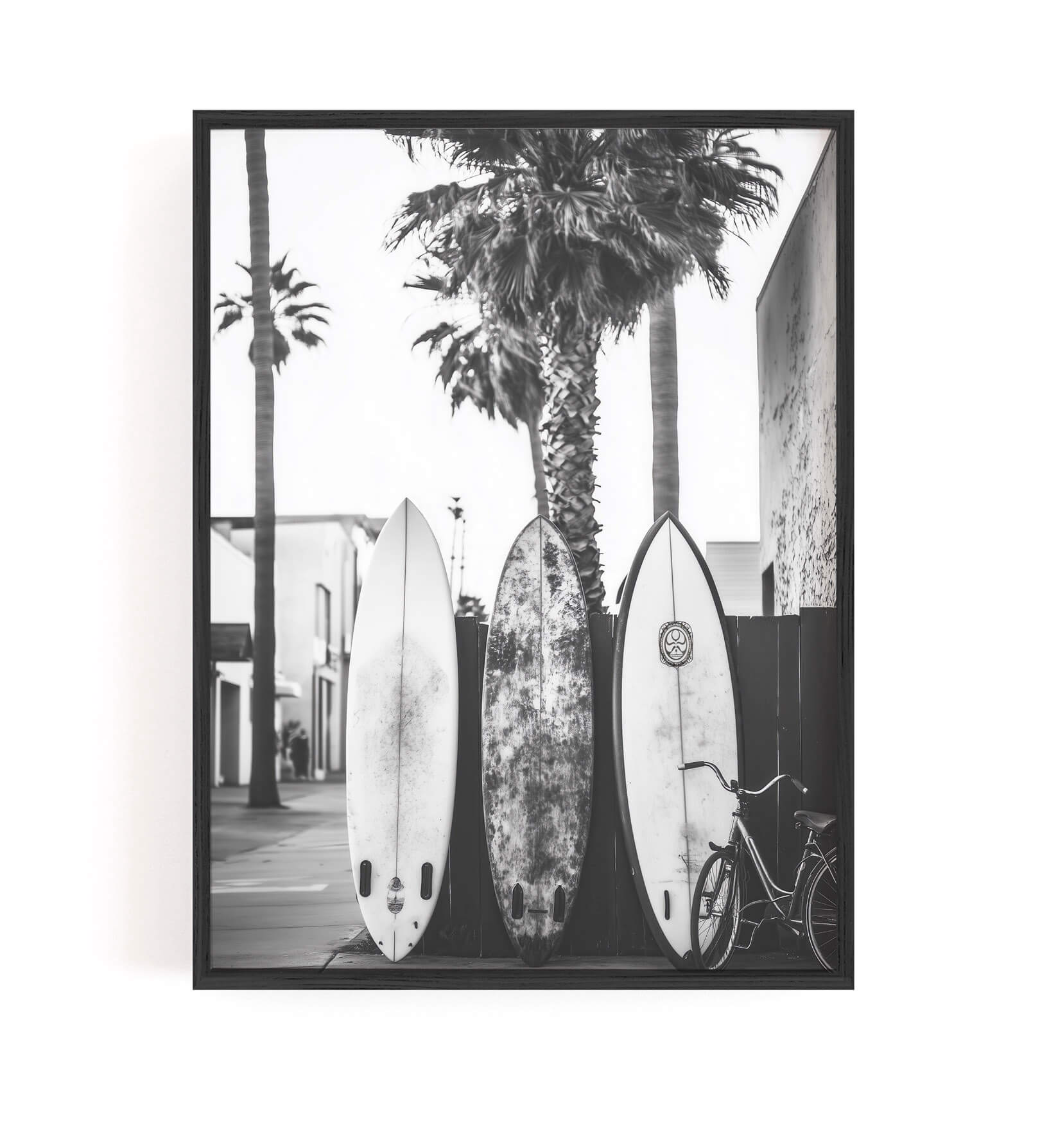 A framed black and white photographic print depicting three surfboards, palm trees, a building, and a bicycle, shown against a plain white background.
