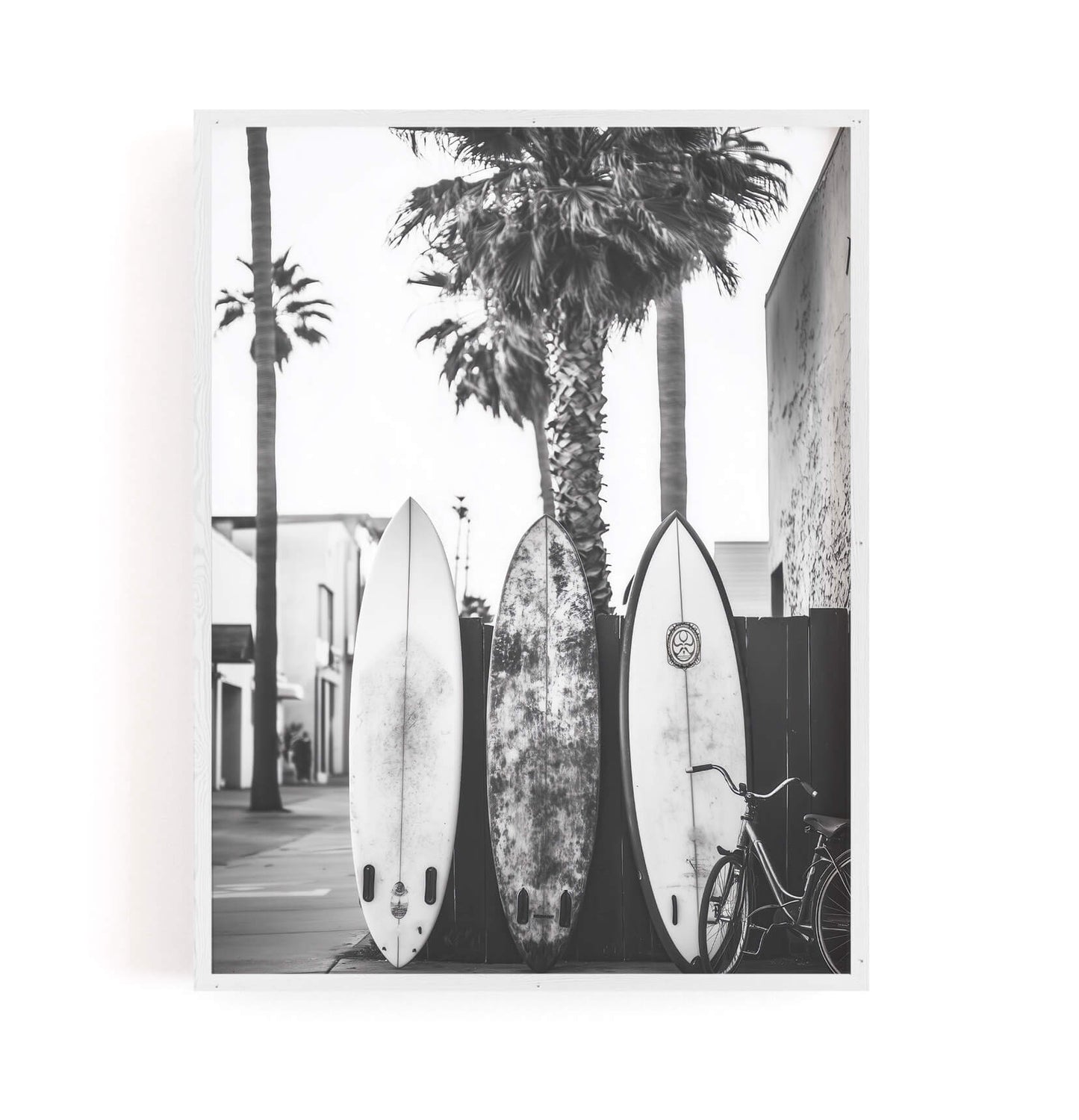 A black and white photographic art print depicting three surfboards leaning against a wall with palm trees and a bicycle, presented as a framed mockup on a white background.