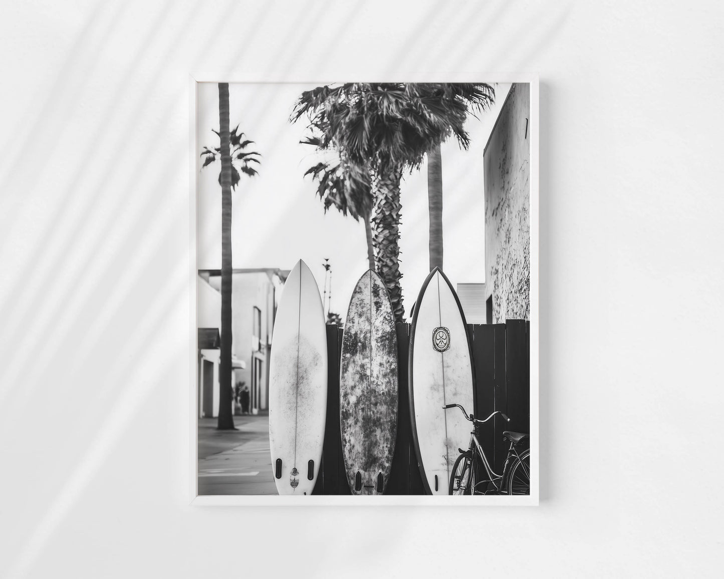 A black and white photographic art print depicting three surfboards leaning against palm trees on a street, displayed on a light gray wall with soft diagonal shadows.