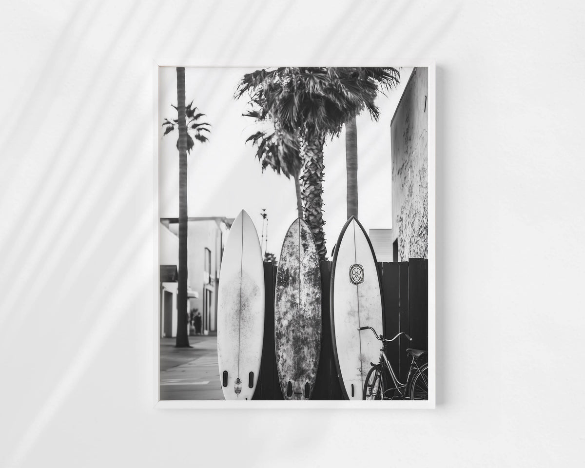 A black and white photographic art print depicting three surfboards leaning against palm trees on a street, displayed on a light gray wall with soft diagonal shadows.