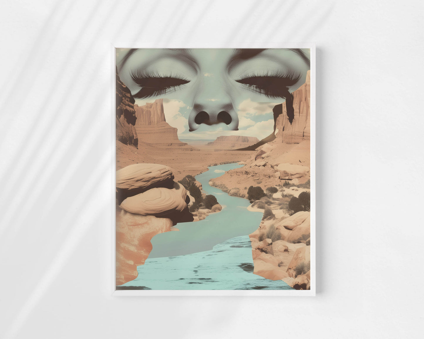 A framed art print featuring a surreal desert canyon landscape with a turquoise river and a large closed-eye face composited across the top, displayed against a light gray background with soft diagonal shadows.