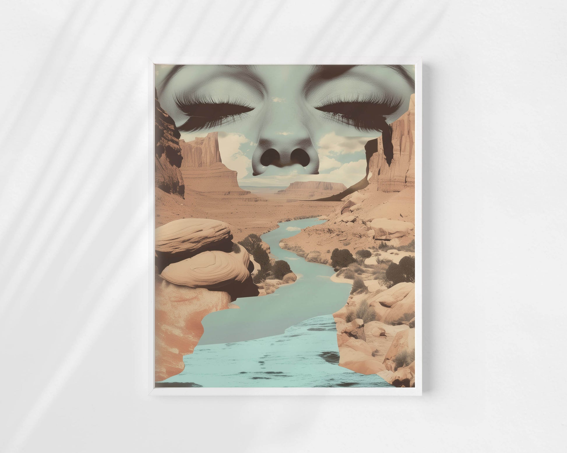 A framed art print featuring a surreal desert canyon landscape with a turquoise river and a large closed-eye face composited across the top, displayed against a light gray background with soft diagonal shadows.
