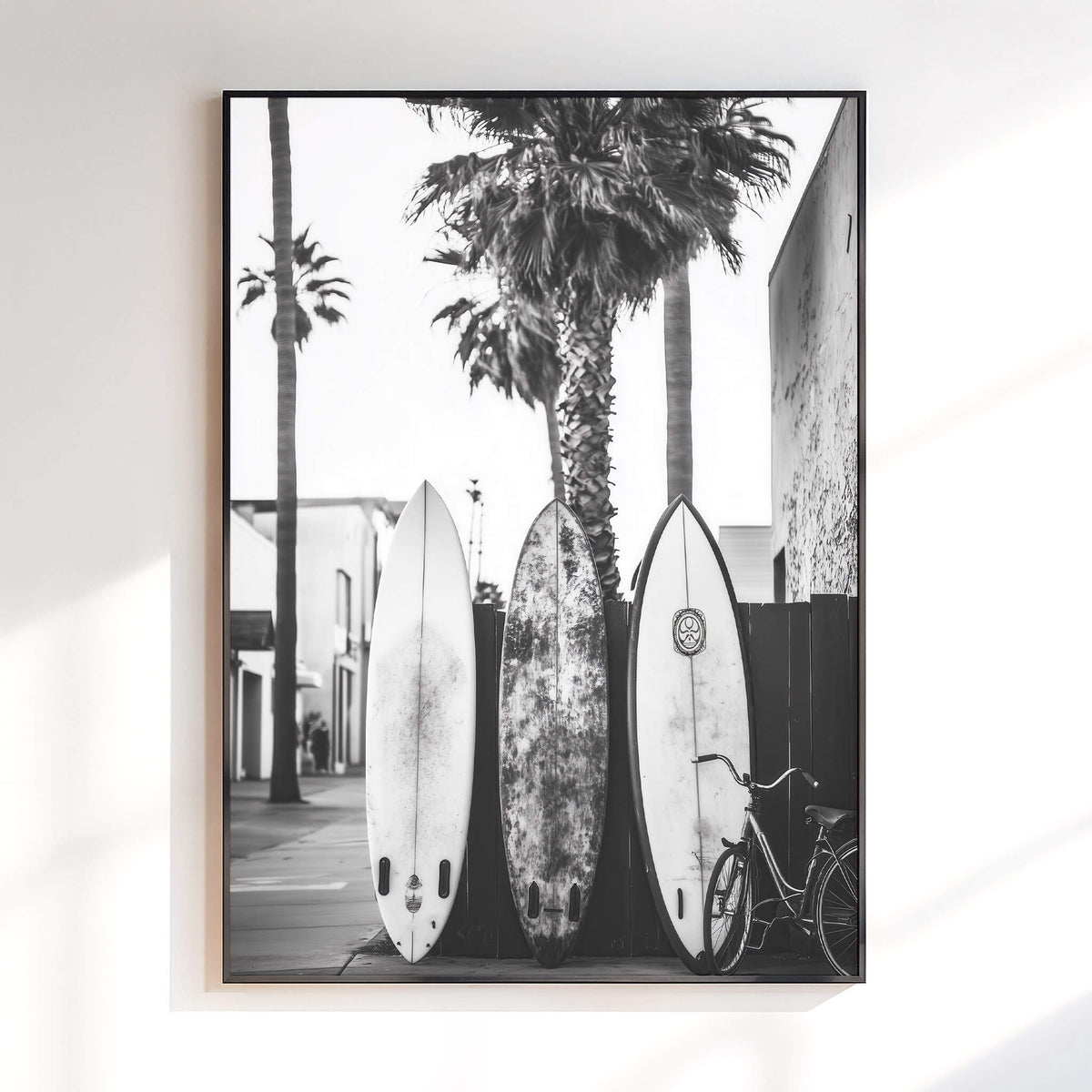 A framed black and white photographic print hanging on a white wall, showing three surfboards leaning against a wall with palm trees and a bicycle in the scene.