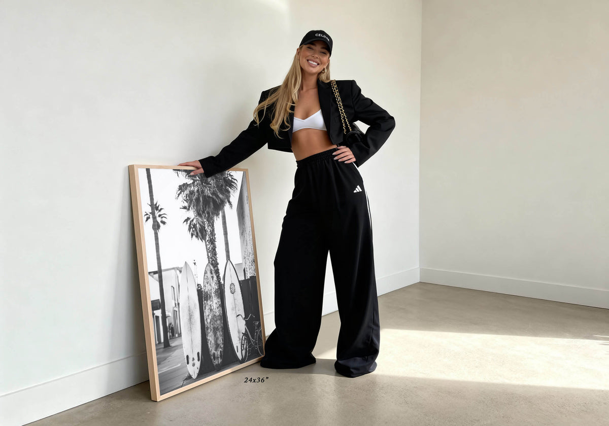 A woman stands in a minimalist room with white walls and concrete floors, leaning against a large framed black-and-white photograph. She is wearing a black cropped blazer, black wide-leg sweatpants, a white sports bra, and a black cap, with a gold chain bag slung over her shoulder.