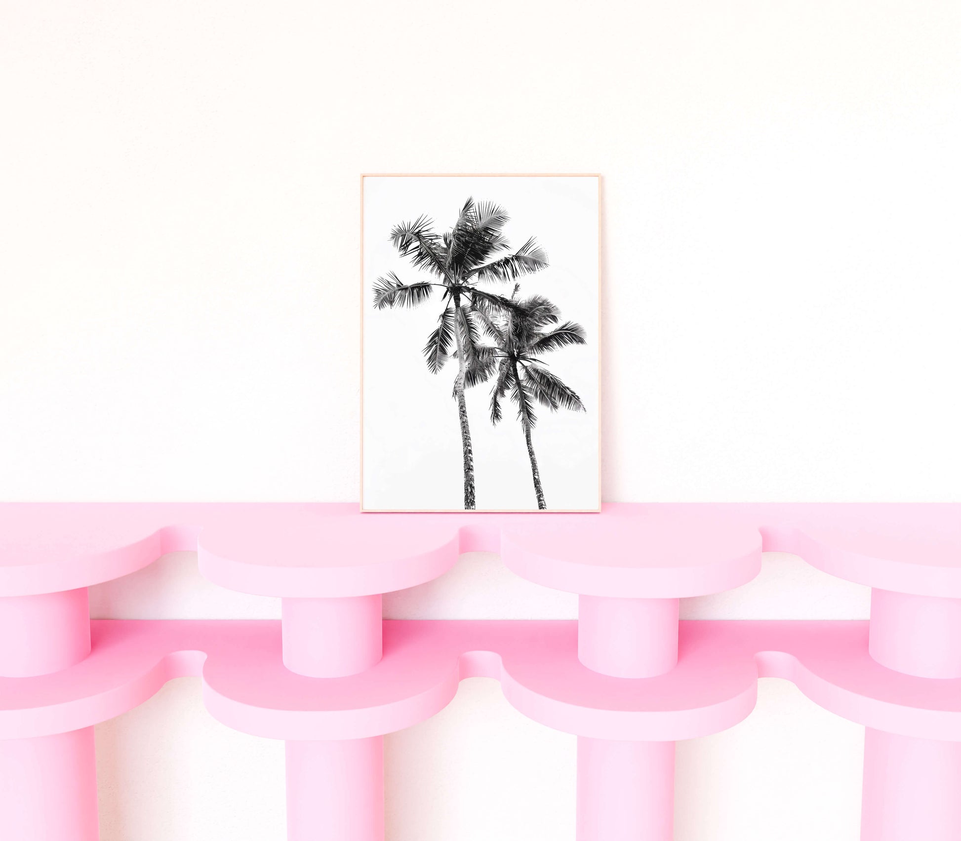 A vertical framed art print featuring a black and white sketch of two palm trees, styled on a pink sculptural shelf against a white wall.
