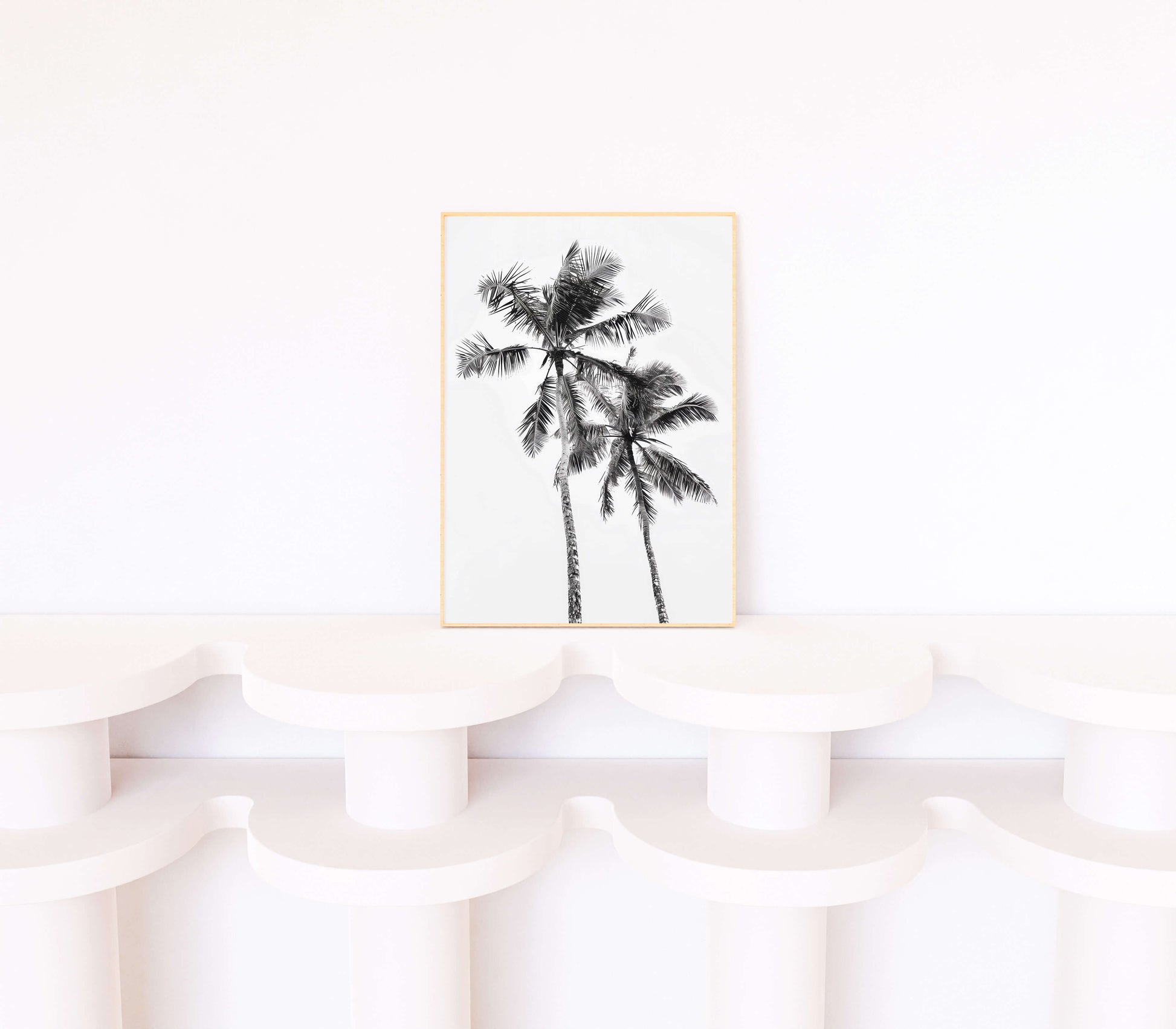 A vertical framed art print featuring a black and white sketch of two palm trees, displayed on a white sculptural shelf against a white wall.