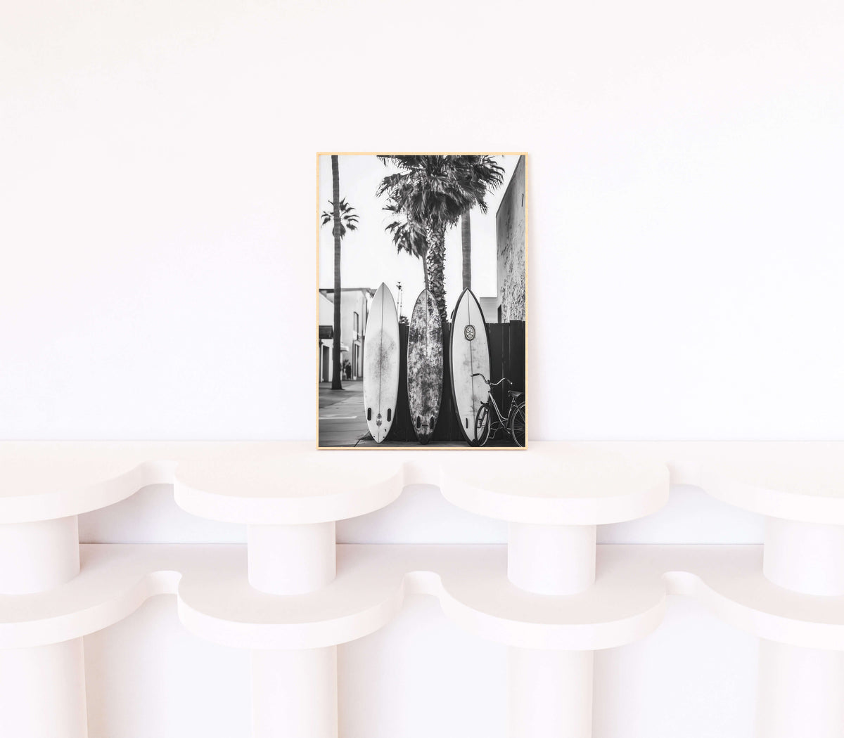 A framed black and white art print featuring palm trees and surfboards, displayed on a white sculptural shelf against a white wall.