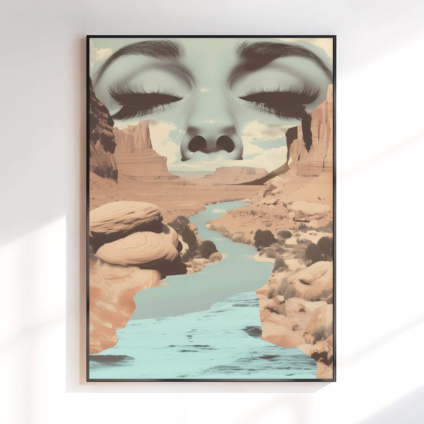A framed wall art print hanging on a white wall, showing a surreal desert canyon landscape with a river and a large face composed of closed eyes and eyelashes blending into the canyon scene.