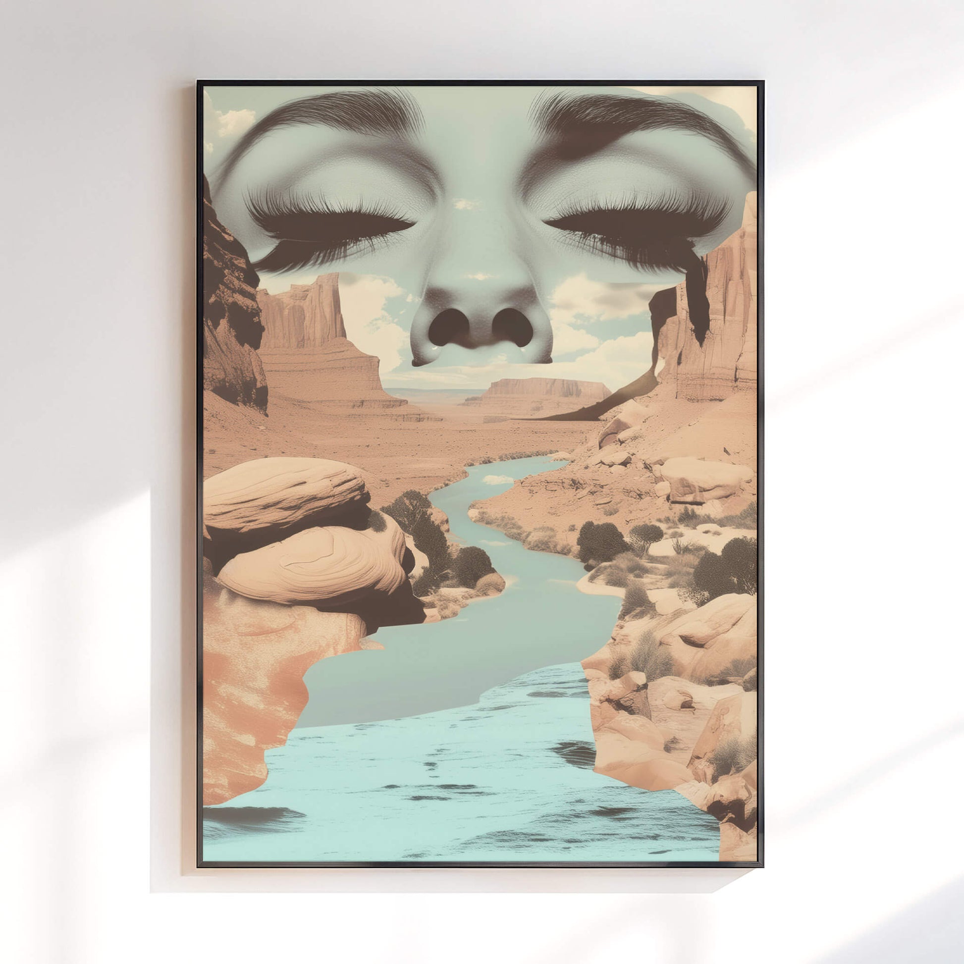 A framed wall art print hanging on a white wall, showing a surreal desert canyon landscape with a river and a large face composed of closed eyes and eyelashes blending into the canyon scene.