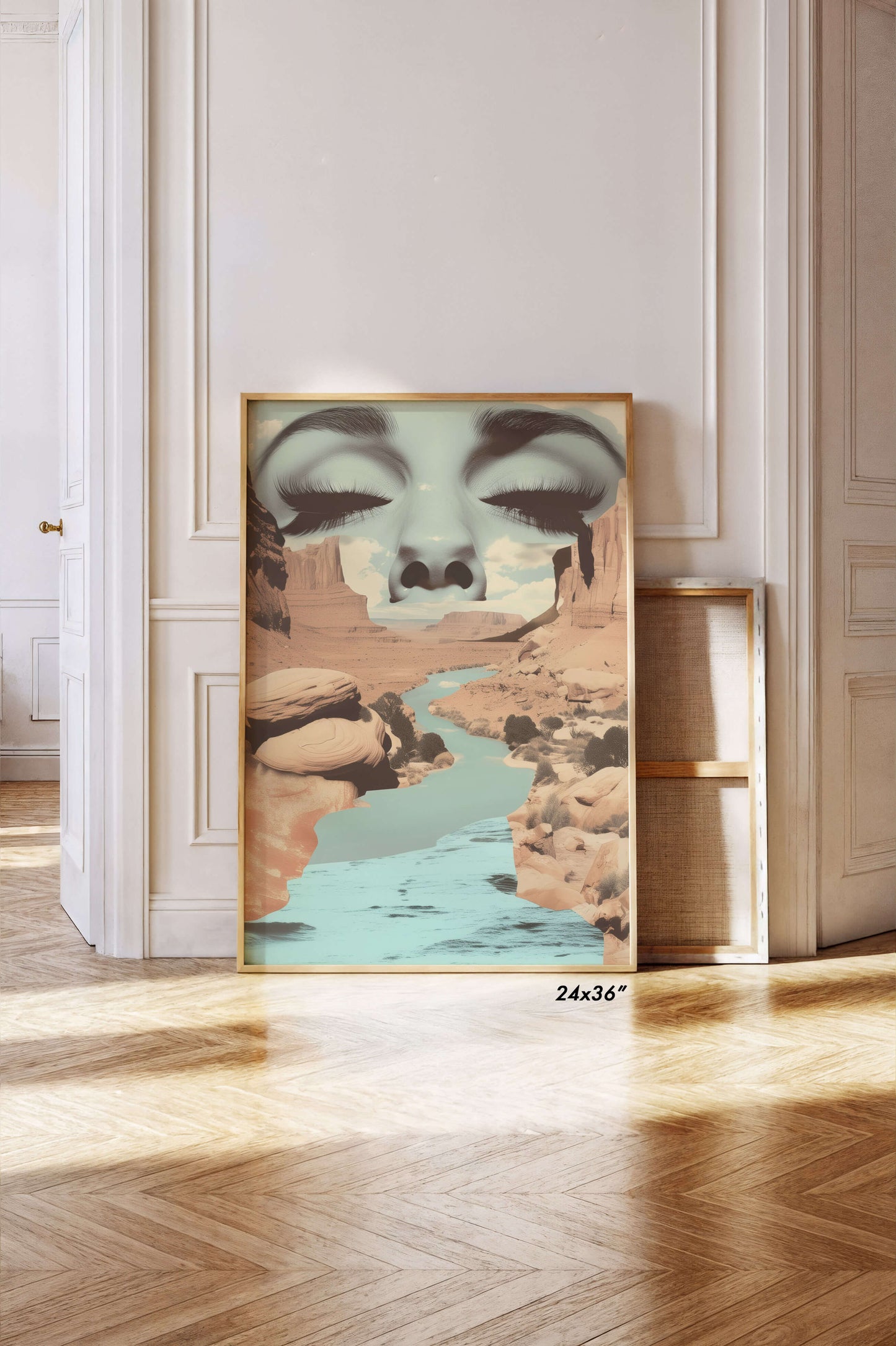 A framed surreal art print leaning against a white paneled wall on a light wood herringbone floor. The artwork features a large stylized face with closed eyes above a canyon landscape with turquoise water.