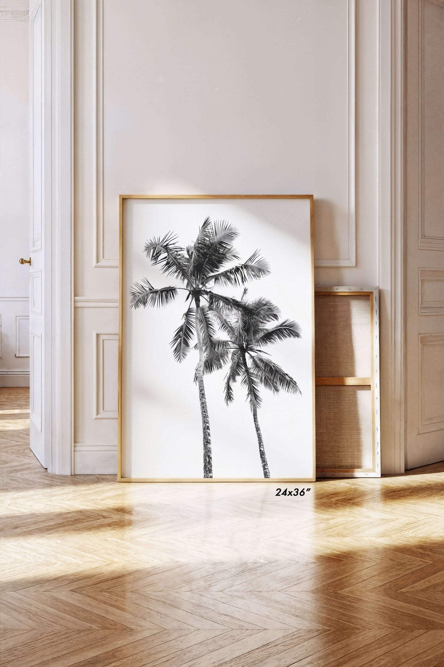 A framed black and white palm tree wall art print leaning against a cream paneled wall on a light wood herringbone floor. The print has a thin gold frame and a small '24x36"' size mark near the bottom right corner.