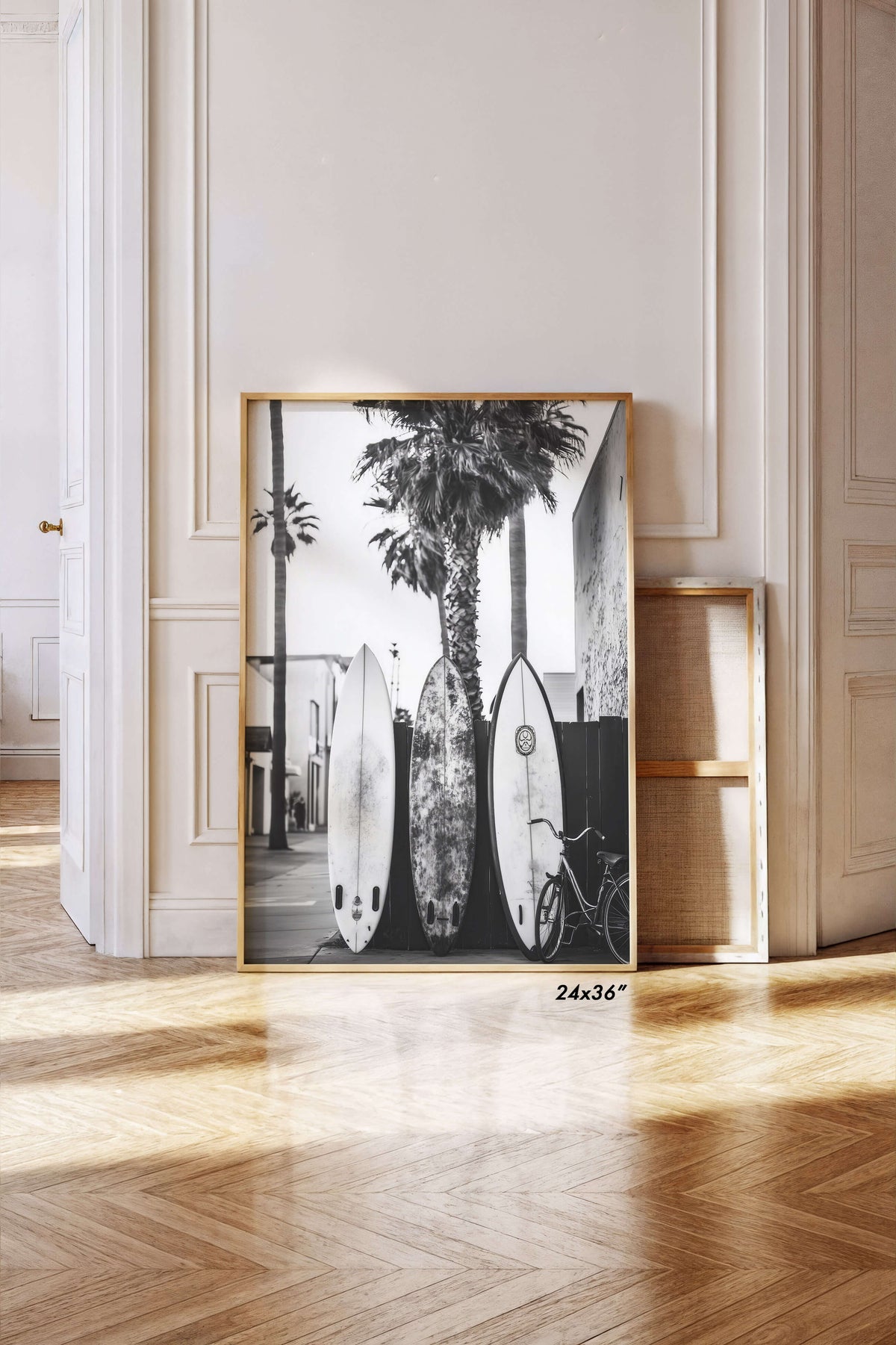 A large framed black and white photographic print leaning against a cream paneled wall on a light wood herringbone floor. The print shows surfboards, palm trees, and a bicycle in a sunlit interior.
