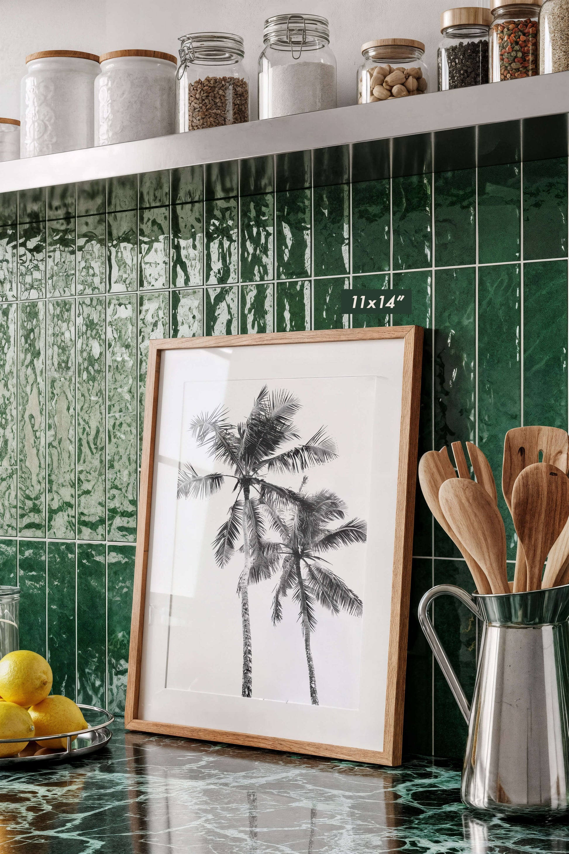 A framed black-and-white palm tree print leaning on a glossy green marble countertop in a kitchen setting with green tile backsplash, glass jars on a shelf, and wooden utensils in a metal pitcher.
