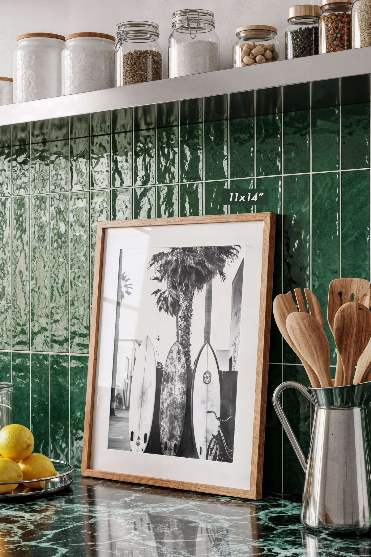 A framed black and white art print featuring surfboards and a palm tree leans against a glossy green tiled kitchen backsplash. The print has a light wood frame and is displayed on a matching green marble countertop next to a metal pitcher of wooden utensils and lemons, with glass jars on a shelf above.