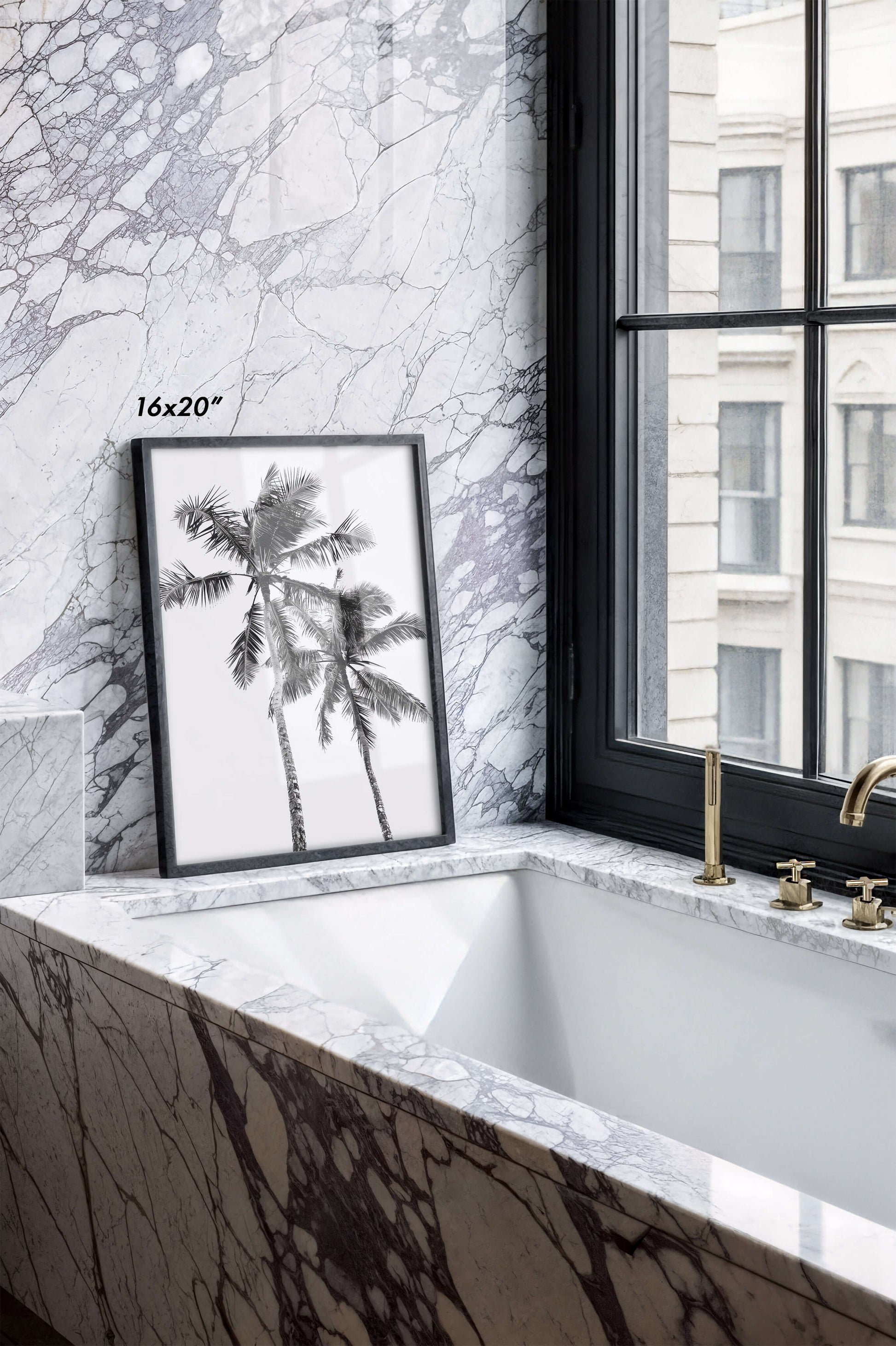 A framed black and white palm tree wall art print leaning against a marble-patterned bathroom wall next to a window with a black frame. The print has a small '16x20"' size overlay.