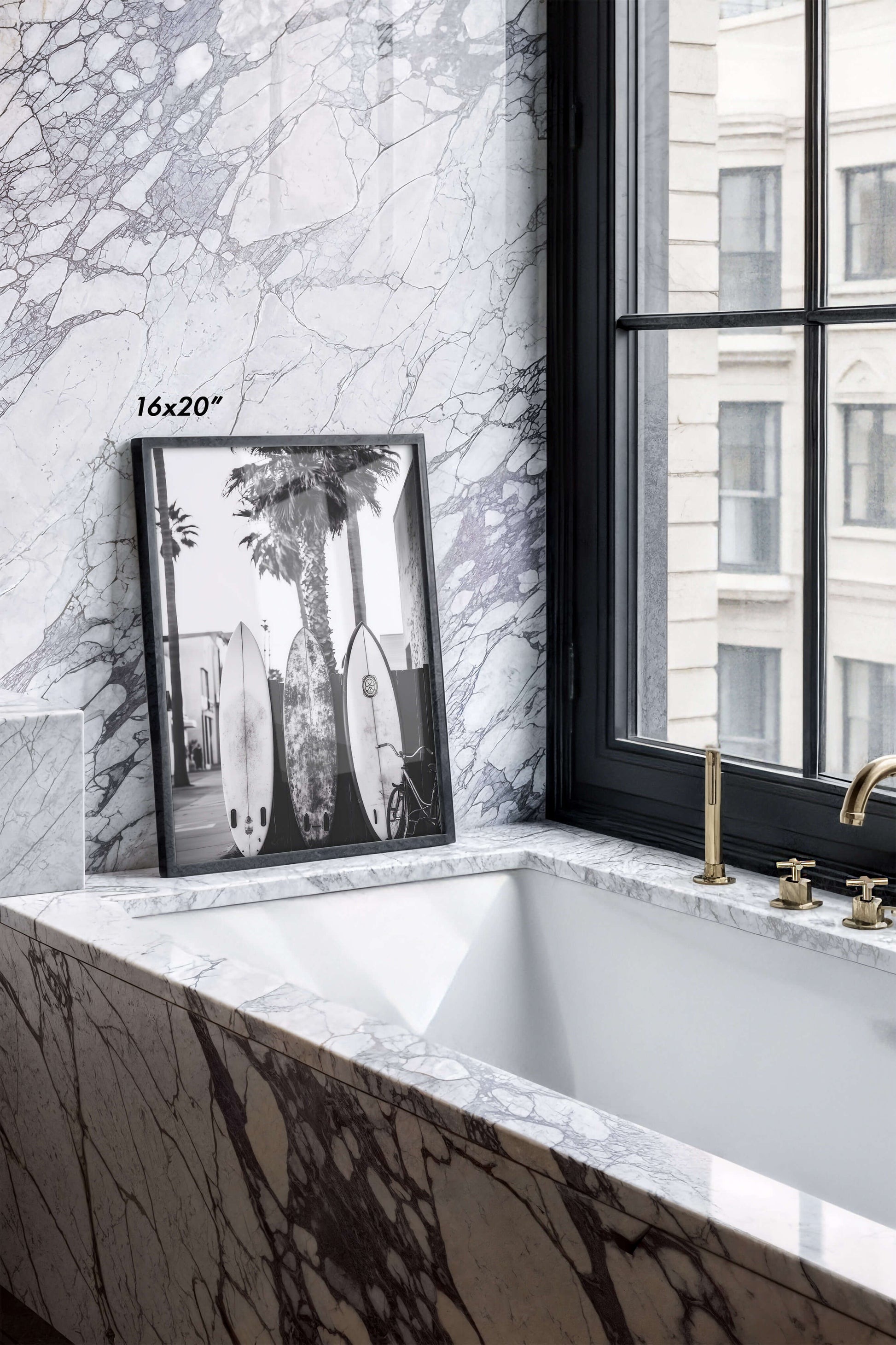 A framed black and white art print leaning against a white marble bathroom wall next to a window. The print features surfboards and a palm tree, with a size label '16x20"' printed near the top left.