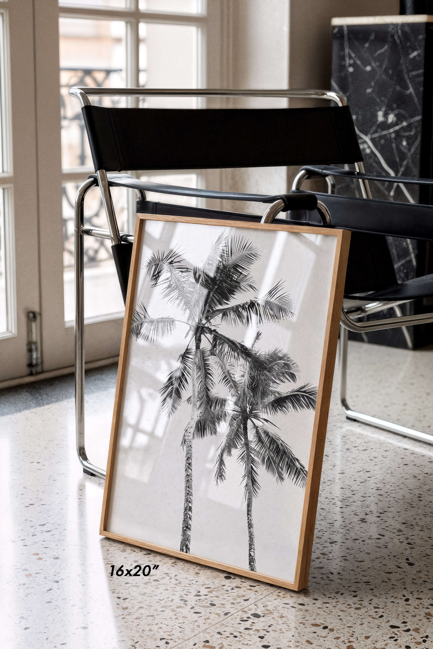 A framed black and white palm tree art print leaning against a black chair on a speckled floor, with a text overlay indicating 16x20 inches.