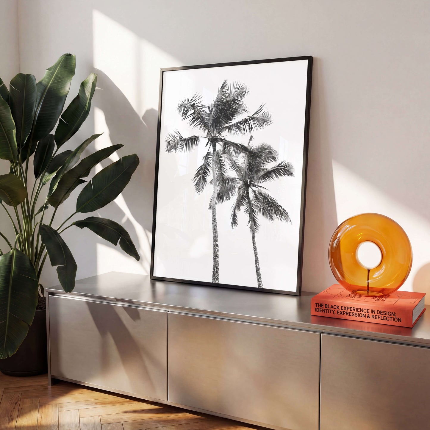 A framed black and white art print of palm trees displayed on a metallic console table, styled in a modern room with a potted plant and an amber glass object.