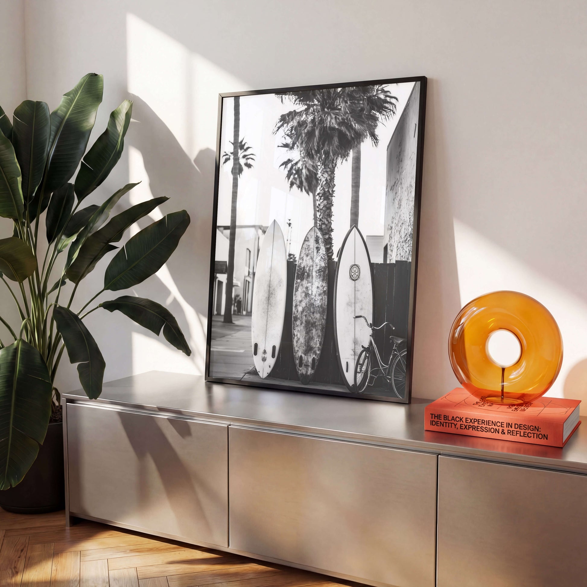 A black and white framed art print featuring surfboards and palm trees, styled on a metallic console table next to a potted plant and an amber glass sculpture.