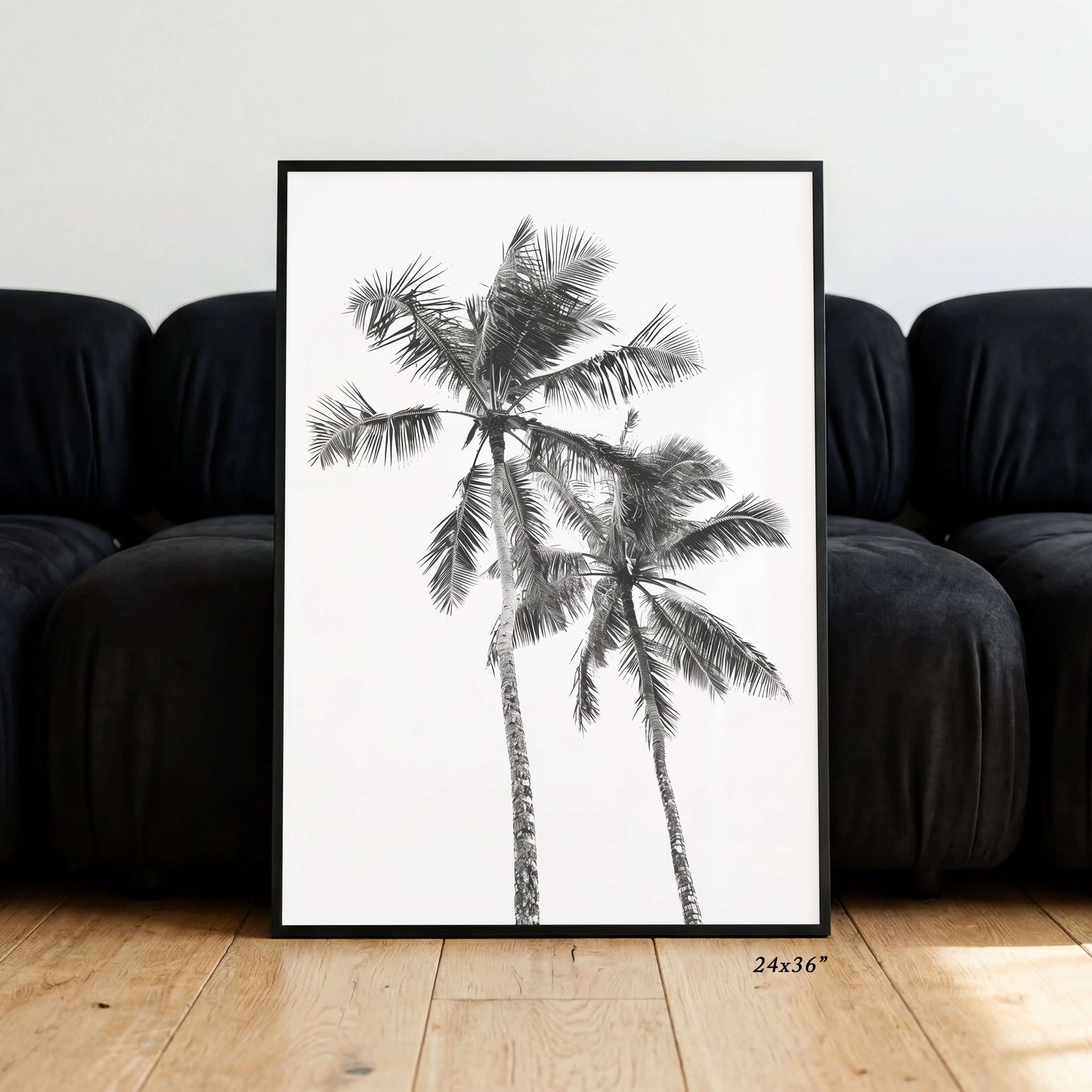 A framed black and white art print of two palm trees leaning against a black sofa on a light wood floor. The print has a white background with detailed black palm fronds and trunks, enclosed in a thin black frame.