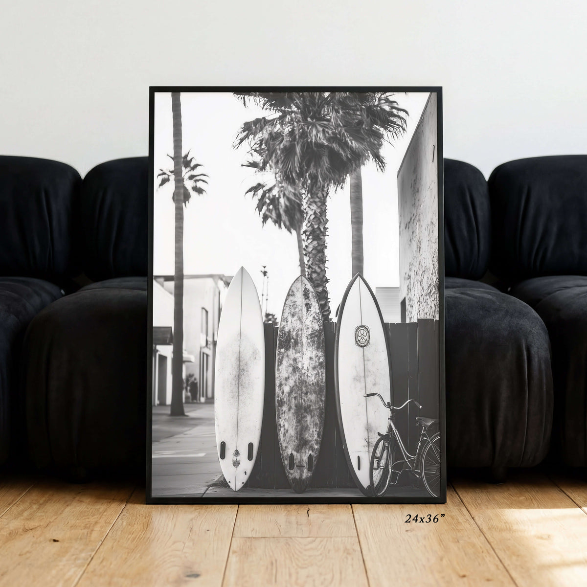 A framed black and white photographic print leaning against a black sofa on a light wood floor. The print shows three surfboards, palm trees, and a bicycle in an outdoor street scene.