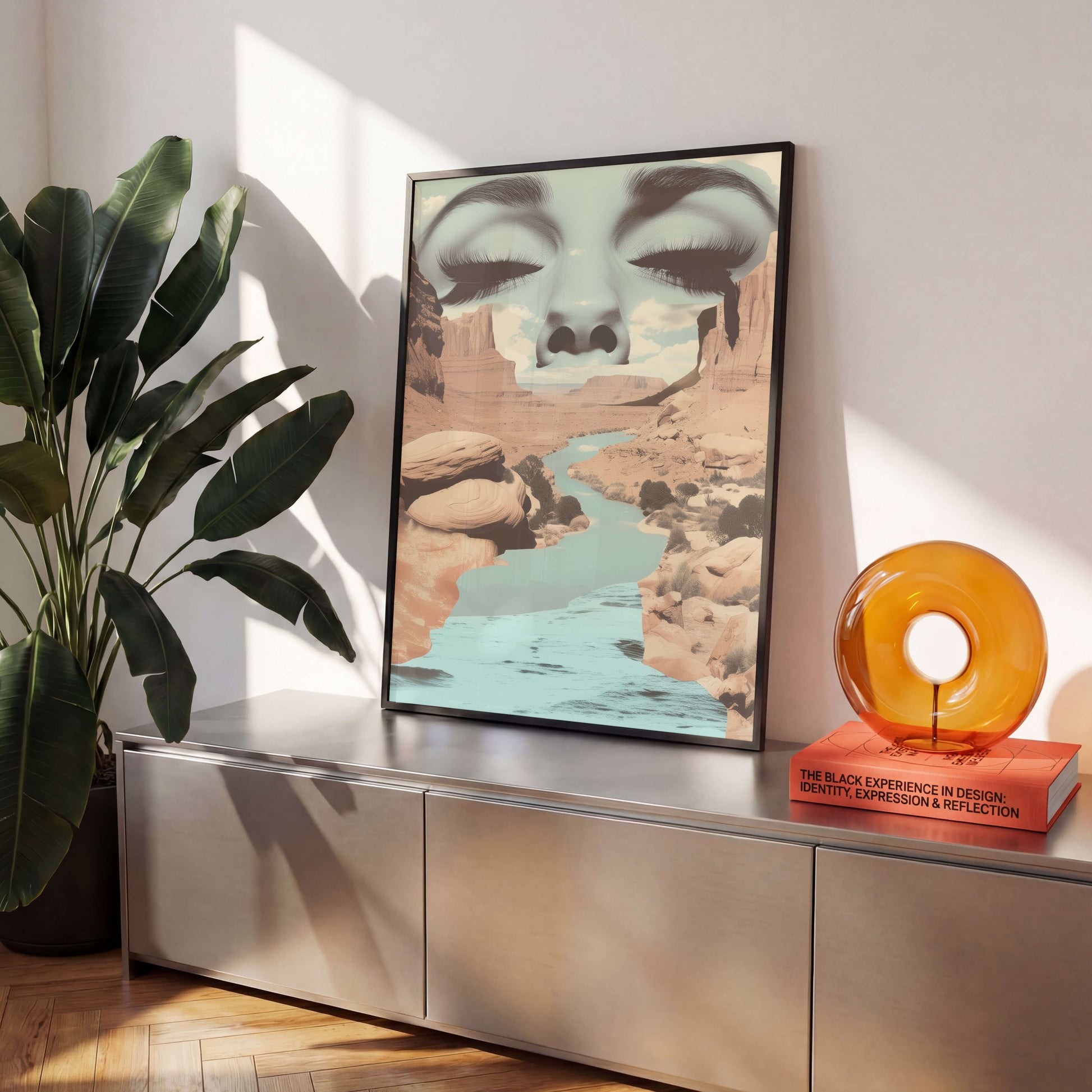 A framed surreal art print featuring a face merging into canyon rock formations above a river, styled on a metallic console table next to a potted plant and an amber glass decor object.