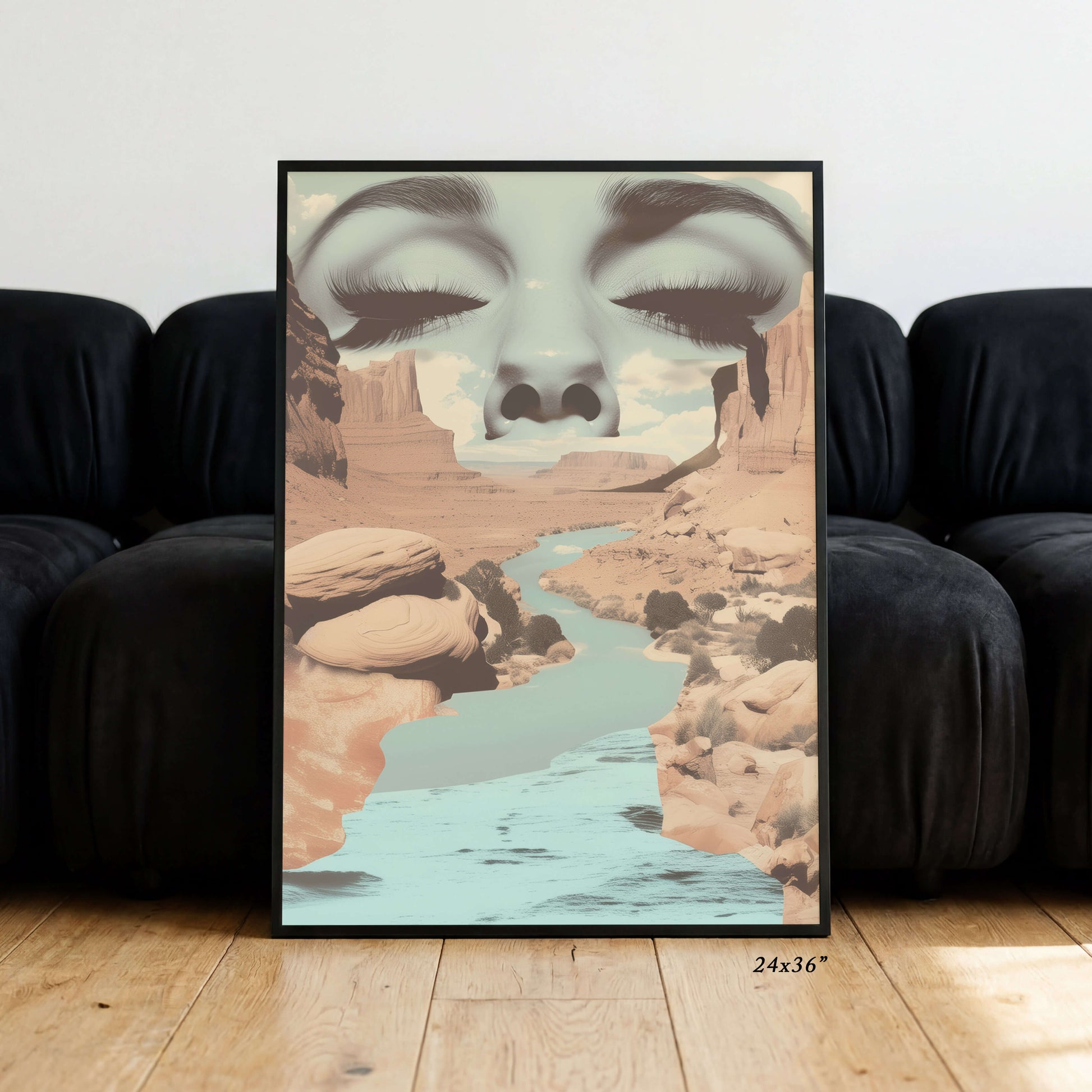 A framed art print leaning against a black couch on a light wood floor. The print features a surreal desert canyon landscape with a river flowing through it, and a large face composed of closed eyes and eyelashes merging into the sky.