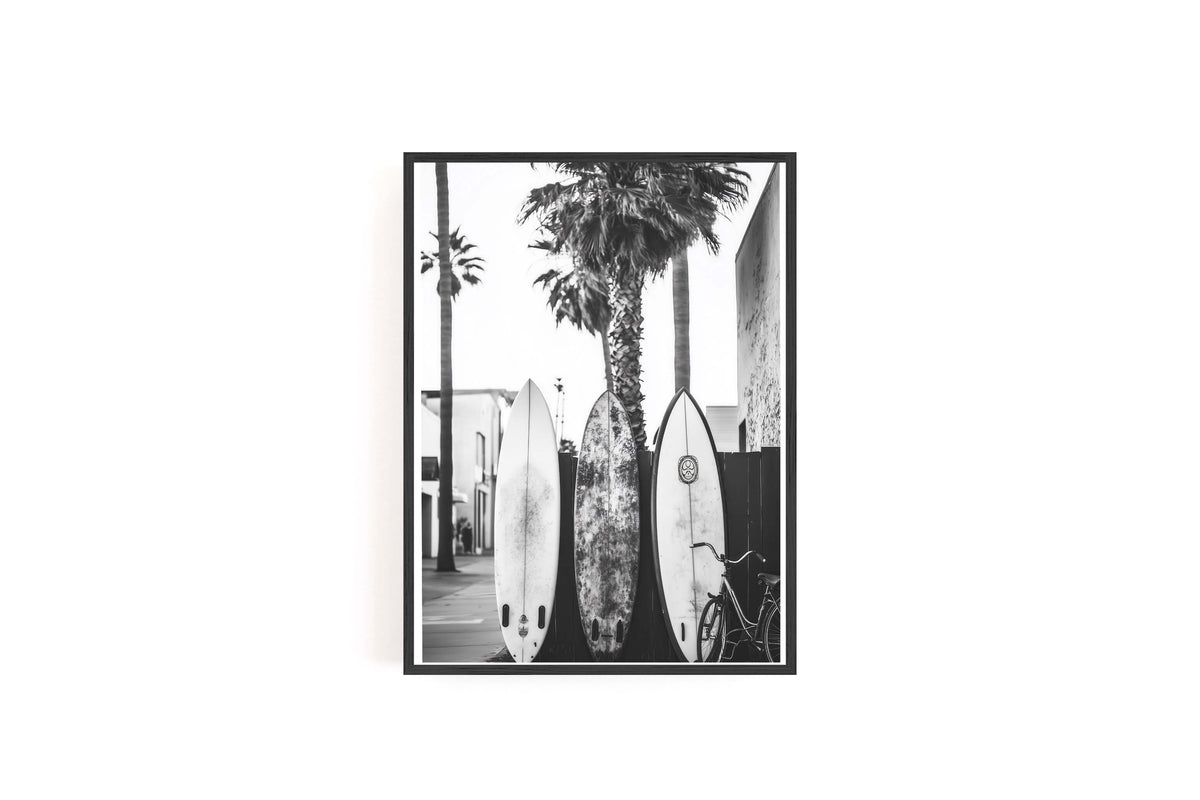 A black and white framed photographic print depicting three surfboards leaning against a wall next to a tall palm tree, shown against a plain white background.