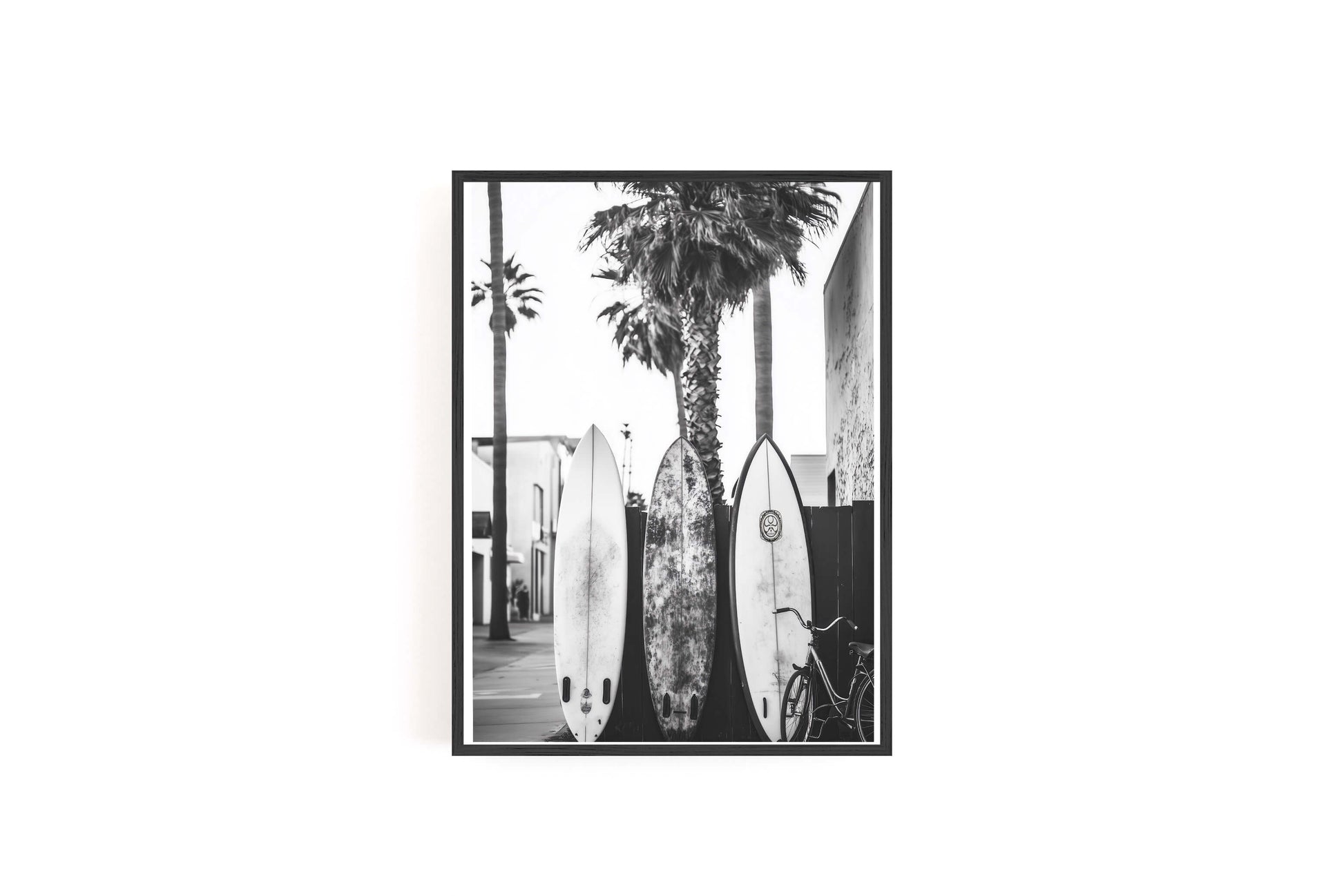 A black and white framed photographic print depicting three surfboards leaning against a wall next to a tall palm tree, shown against a plain white background.