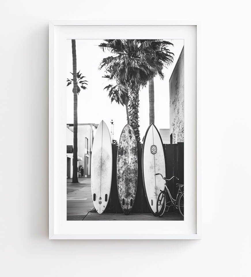 A framed black and white photographic print of three surfboards leaning against a palm tree on a street, shown hanging on a white wall.