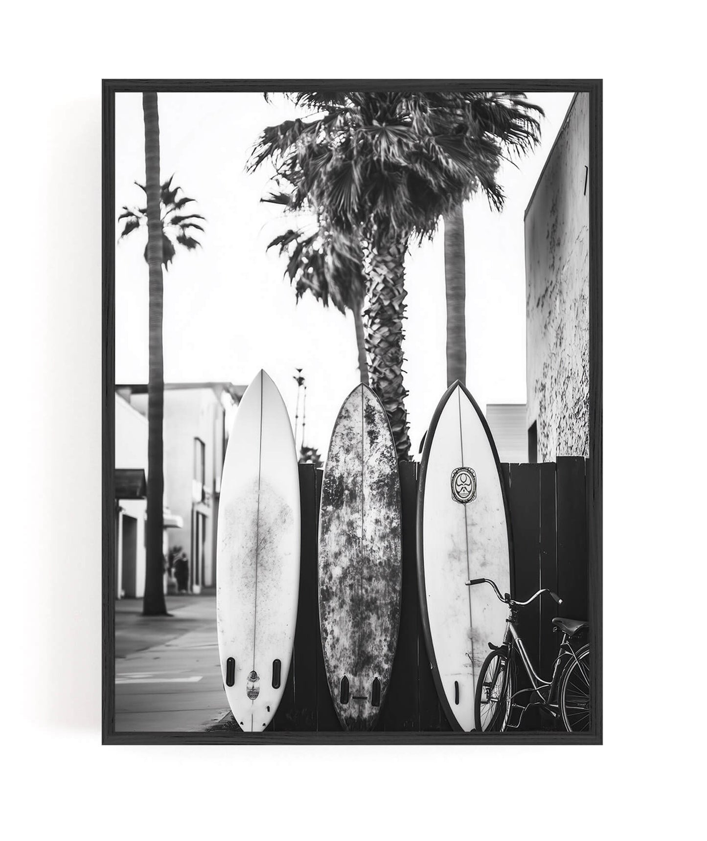 A framed black and white photographic print featuring three surfboards leaning against a palm tree on a street, with a bicycle at the right and buildings in the background.