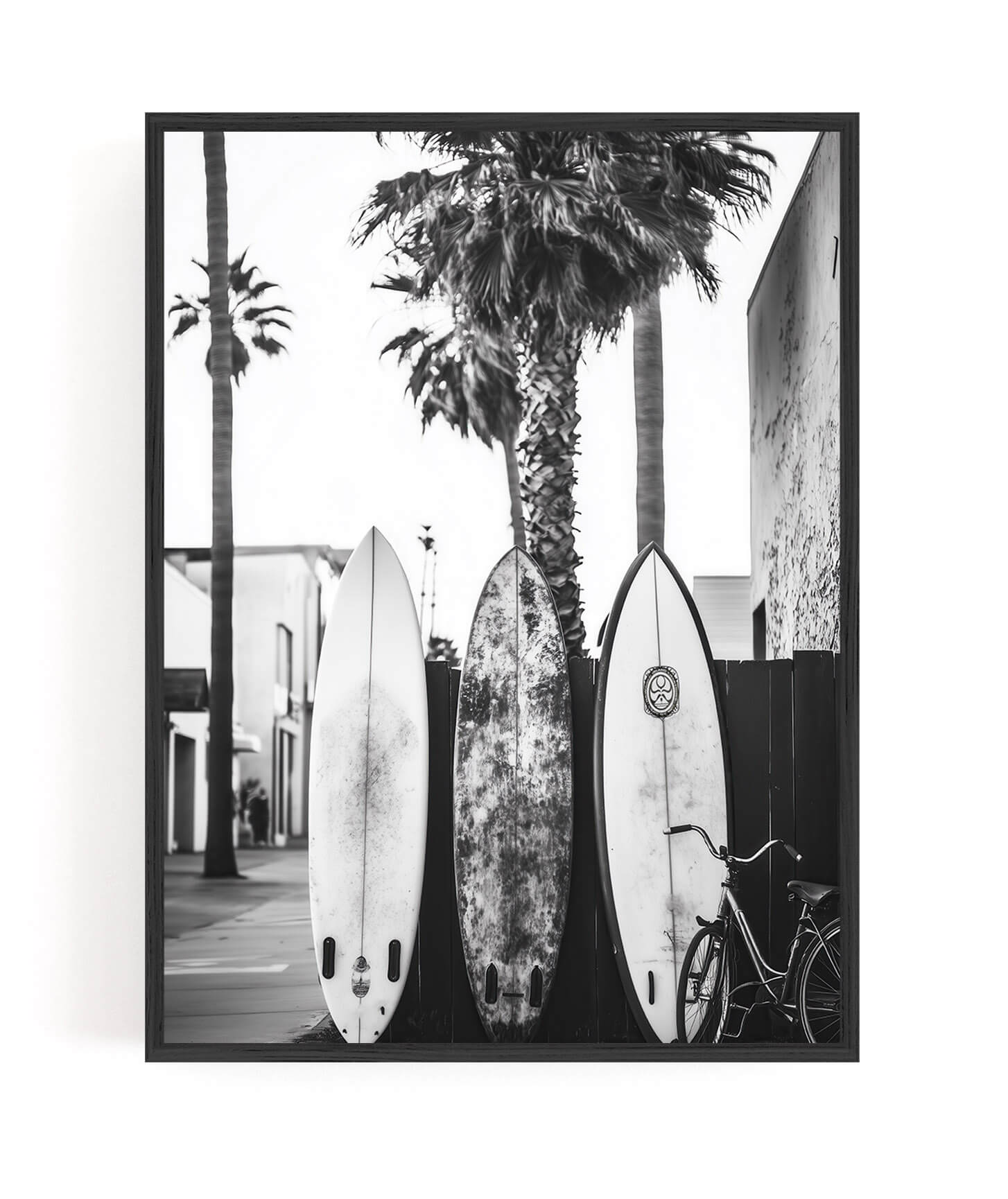 A framed black and white photographic print featuring three surfboards leaning against a palm tree on a street, with a bicycle at the right and buildings in the background.