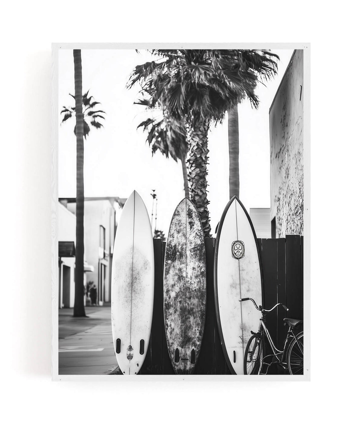 A black and white photographic art print depicting three surfboards leaning against a wall with palm trees and a bicycle, presented as a framed mockup on a white background.