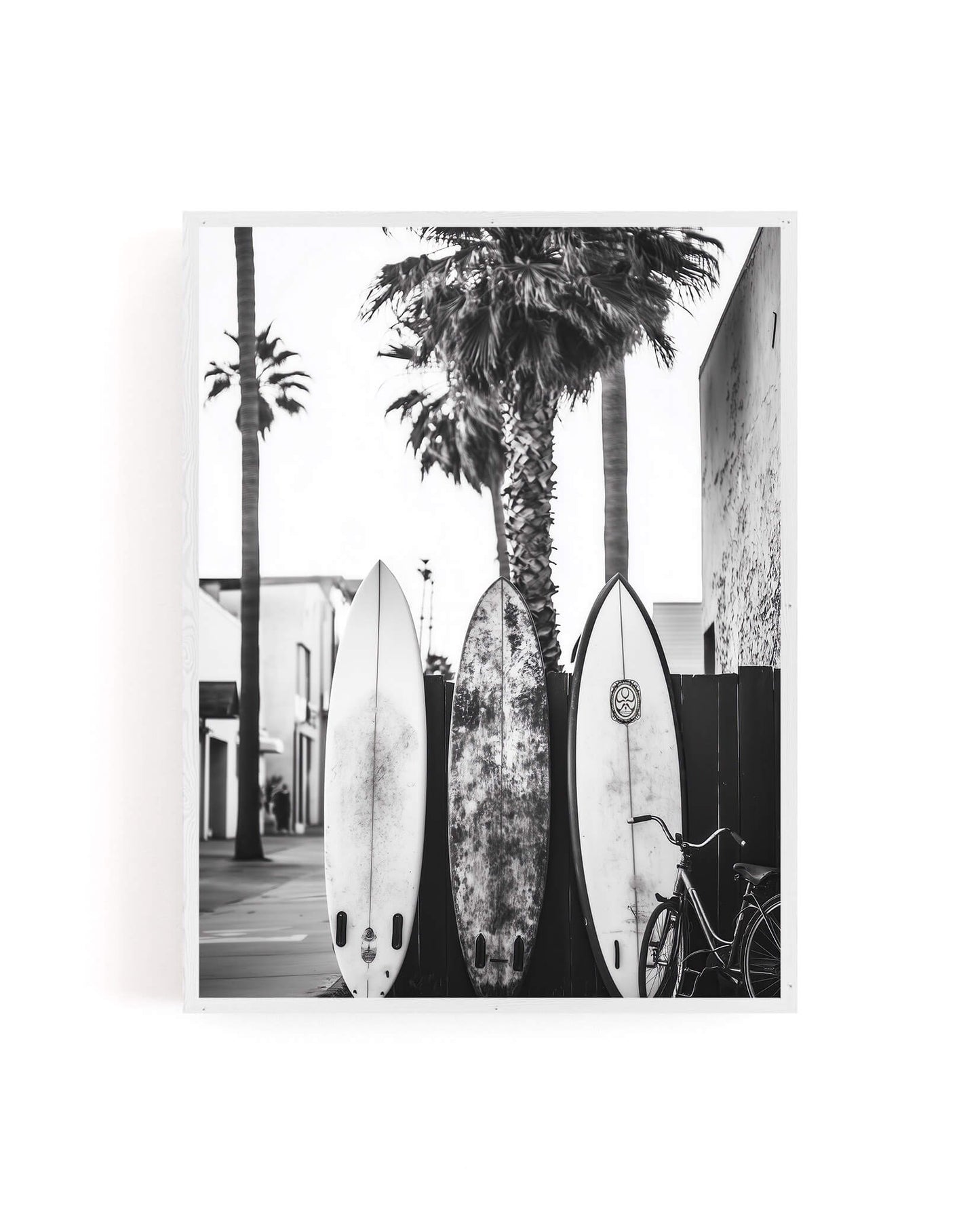 A black and white photographic art print depicting three surfboards leaning against a wall next to palm trees and a bicycle, shown as a square canvas with a white border on a white background.